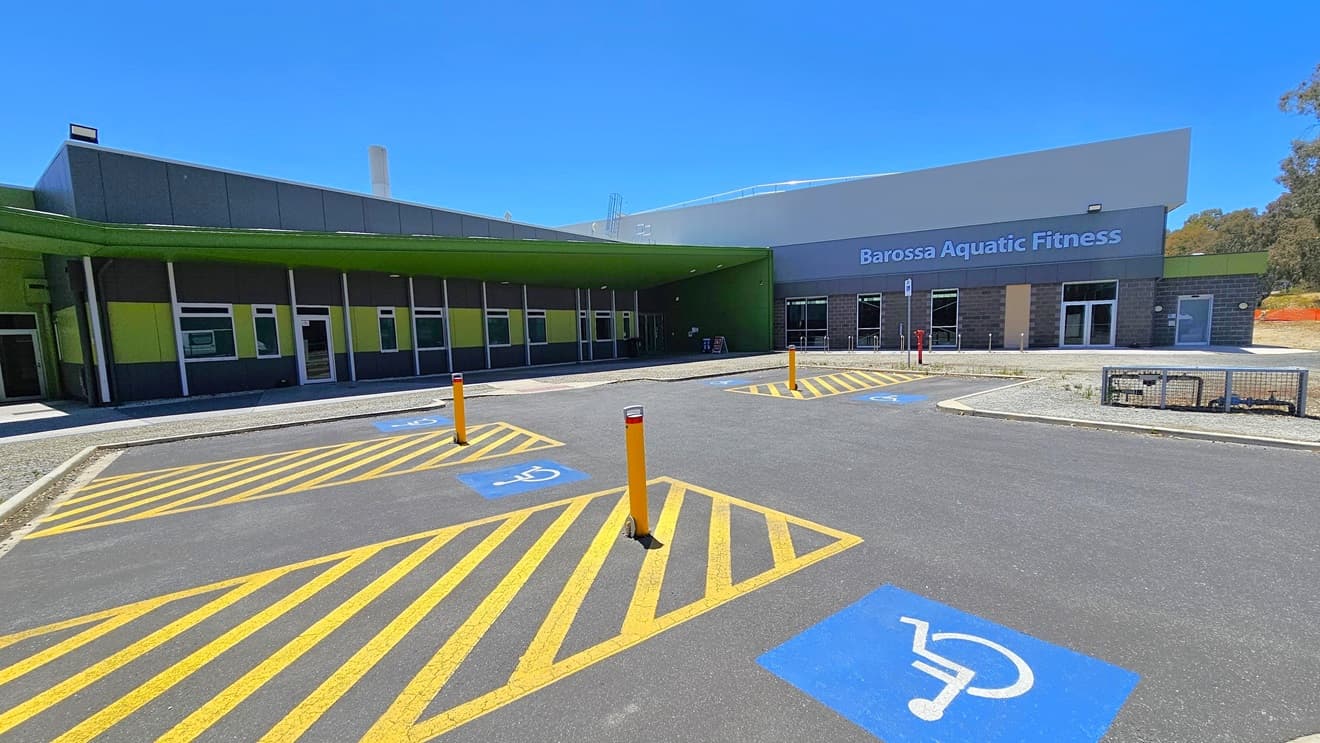 There are five accessible parking spaces near the entrance, each clearly marked with blue wheelchairs and yellow diagonal markings. Yellow bollards are also in place. The building on the right has alternating colors of green and grey. To the right is the entrance, featuring a huge "Barossa Aquatic Fitness" sign on the wall. A barricaded section with a water meter and controls is situated on the right.