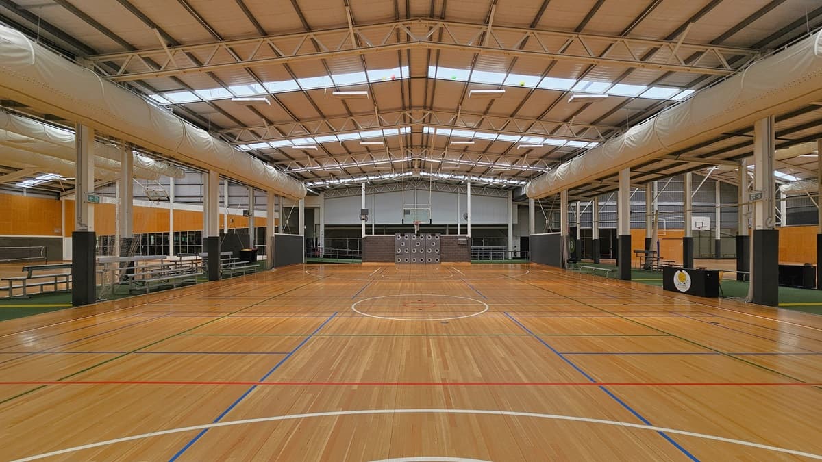 The court has a spacious area with timber floors marked for ball games. The ceiling has an industrial look with exposed beams, large air conditioning vents, fluorescent lights, and natural skylights. On the left, there is an area with green carpets and tiered seating. On the right, another area with green carpets is equipped with bench chairs and tables. The surrounding wall has a wooden design.