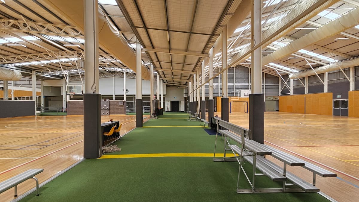 In the centre is an area with green carpets marked with yellow lines. Tiered seating is arranged on both sides, along with tables and yellow plastic chairs. On both sides, there are basketball courts with timber flooring marked for ball games. The ceiling has an industrial look with exposed beams, large air conditioning vents, fluorescent lights, and natural skylights. The surrounding wall features a wooden design.