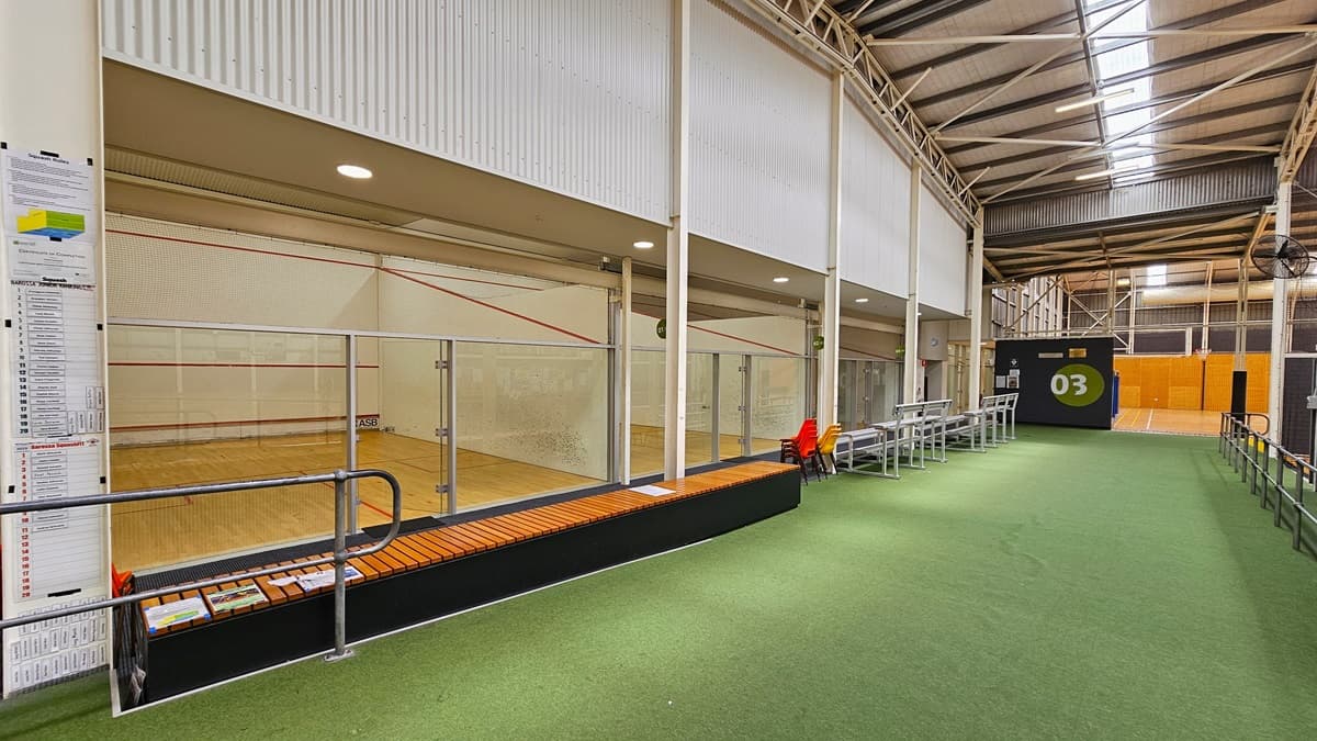 The squash court features a timber floor area with red markings, enclosed by a net and stainless steel barriers. On the left, there is a large map showing the vicinity. The middle is carpeted in green, with stainless tiered seating on the left, accompanied by red and yellow plastic chairs. On the right, metal rails are visible. The wall is painted white with a corrugated design, and the roof has an industrial look with exposed beams, natural skylights, and fluorescent lights.