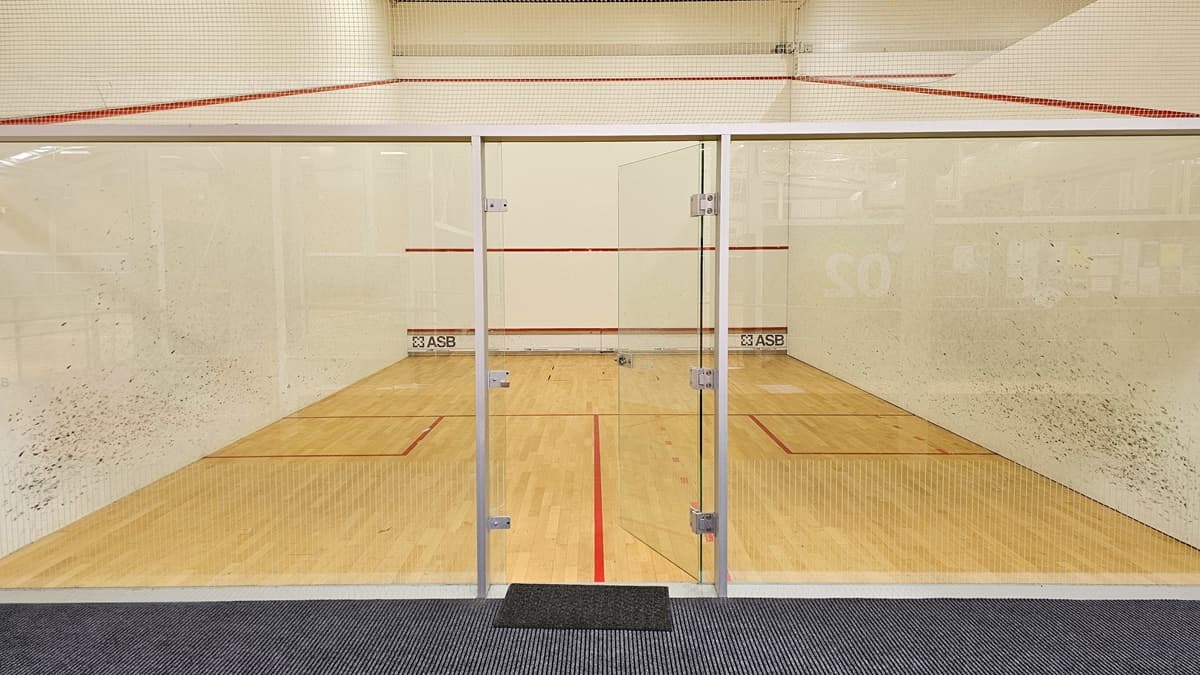 The squash court has a timber floor with red markings, enclosed by a net and stainless steel barriers. White walls with black markings surround the court, and there is a glass door in the middle that opens inward, serving as the entry point. The middle wall features an inscription "ASB" on the lower section.