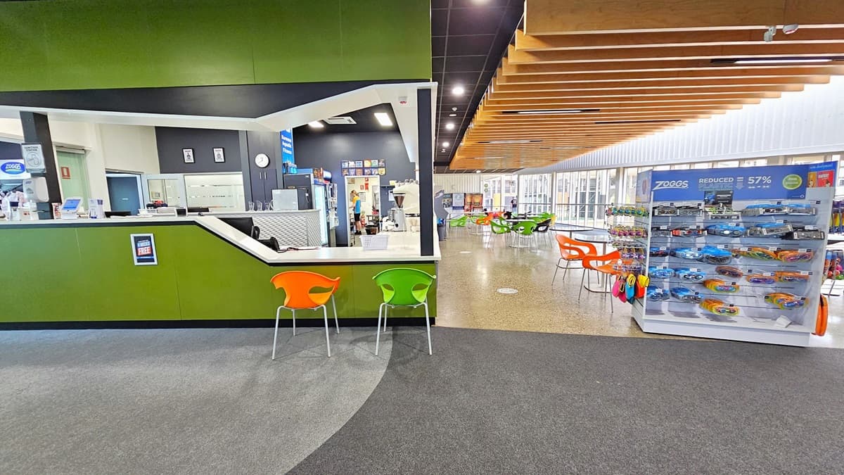 To the left is the reception with green walls and a brown ceiling. The reception counter has a sloping design for seated assistance. An alcohol dispenser and a card-swiping machine are placed on the counter. Orange and yellow-green plastic chairs are positioned for seating. A display rack with goggles and swimming accessories is on the right. Movable plastic tables are placed in front of the cafe. The floor is tiled with granite, and the ceiling has a timber wooden panel design.