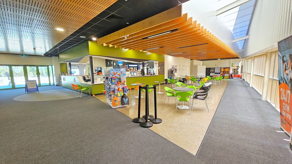 To the left is the entrance and the reception. The reception counter has a sloping design for seated assistance. Orange and yellow-green plastic chairs are positioned for seating. On the right, there is a display rack with goggles and swimming accessories. Movable plastic tables are placed in front of the cafe. The floor is tiled with granite, and the ceiling has a timber wooden panel design. Ribbon bollards are also in place. A large banner on the right advertises the gym's 24-hour operations.