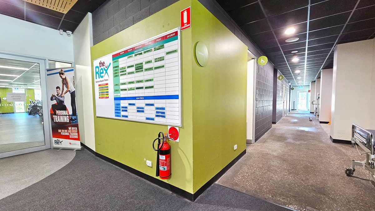 On the left is the entrance to the gymnasium with a green wall displaying The Rex schedule and a fire extinguisher. A gym banner is positioned before the door. To the right is a lobby leading to accessible bathrooms and other amenities. Circular lights on the grey ceiling illuminate the area.