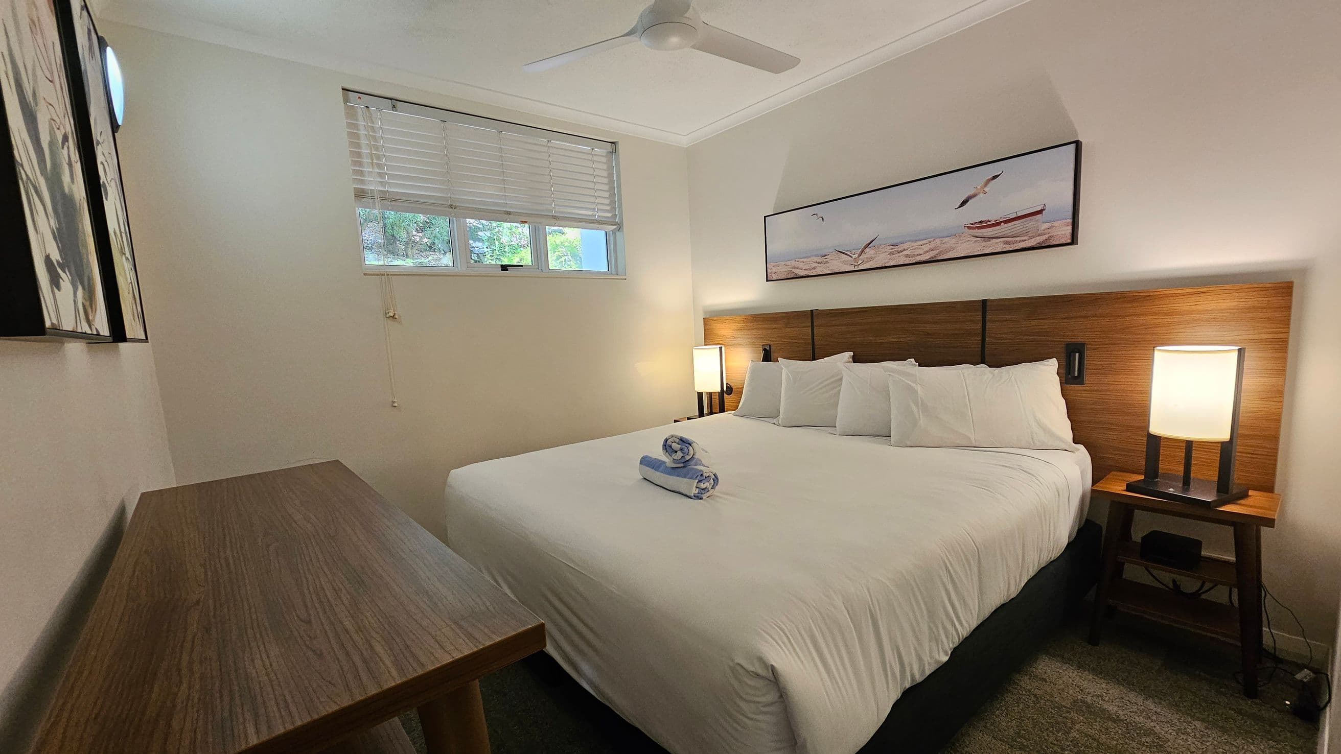 A bedroom with a large bed in the centre, topped with white linens and a rolled towel. On the right, a wooden headboard with a bedside table and lamp. Above the bed, a framed picture of a beach scene. On the left, a wooden desk and a piece of artwork on the wall. A window with blinds is on the back wall, allowing natural light. The ceiling has a white fan. The floor is carpeted.