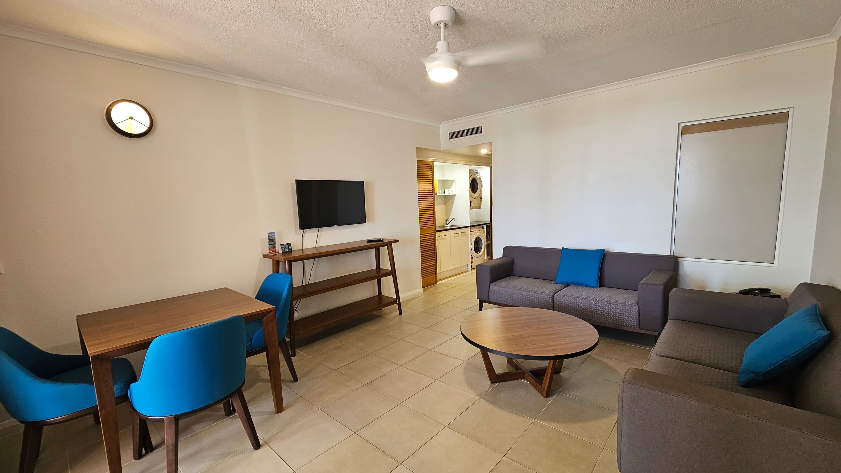 A living room with a grey sofa and blue cushions on the right, and a round wooden coffee table in front. On the left, there's a wooden dining table with blue chairs. A wall-mounted TV is above a wooden shelf unit. In the background, a doorway leads to a laundry area with a washing machine and dryer. The floor is tiled, and a ceiling fan is visible. A small wall clock is on the left wall.