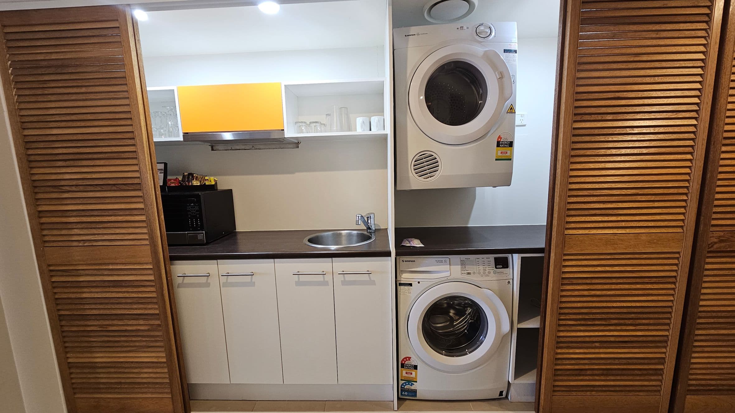 A compact laundry and kitchenette area is enclosed by wooden louvre doors. On the left, a black microwave sits on a dark countertop above white cabinets. A small stainless steel sink is centrally located. Above, open shelves hold glasses and cups, with a yellow panel adding colour. On the right, a white dryer is mounted above a washing machine. The floor is tiled, and the ceiling has recessed lighting.