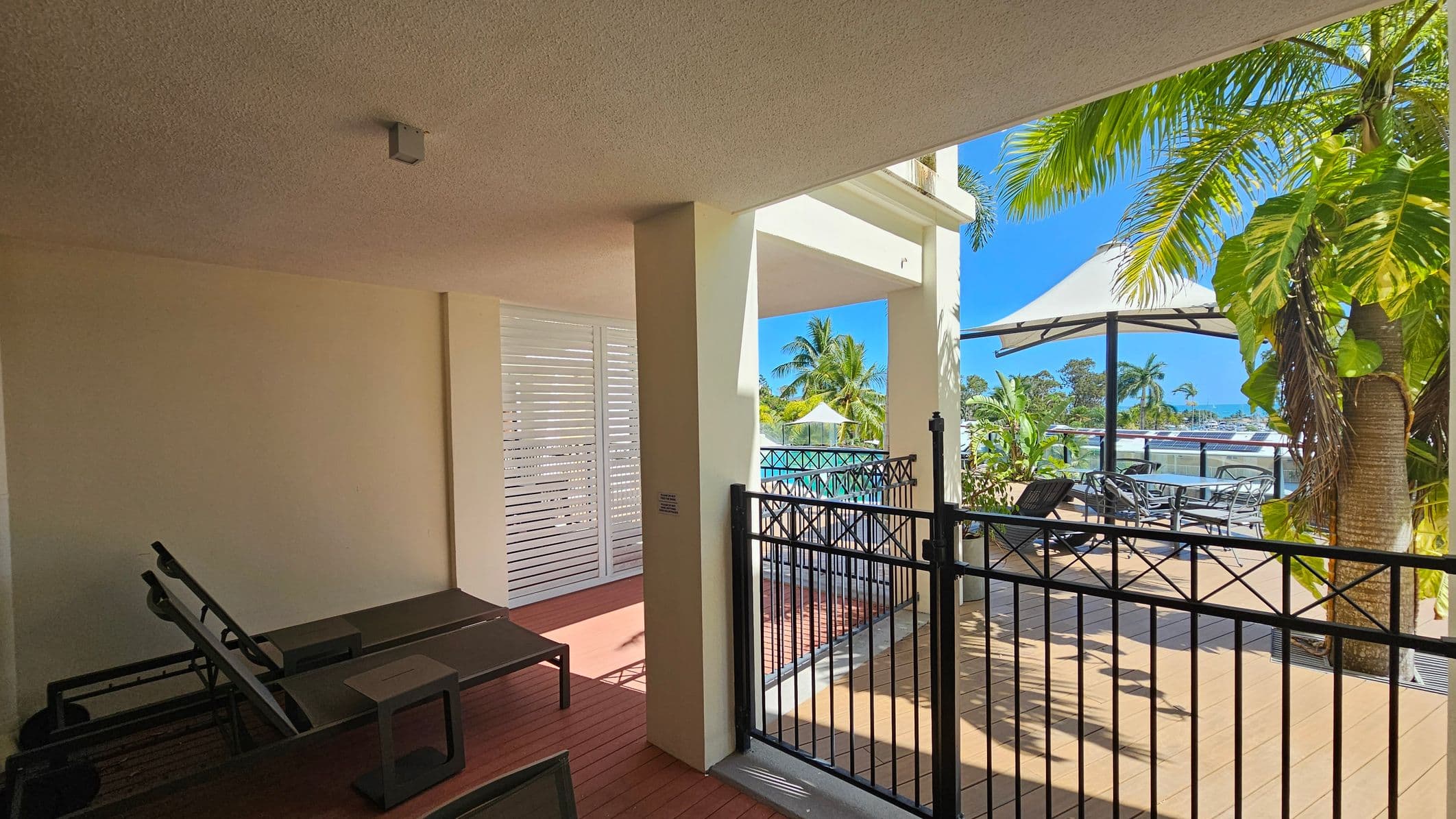 A covered patio area with two dark sun loungers and a small table on the left. The floor is wooden decking. In the centre, a white louvred door and a supporting column. On the right, a black metal gate opens to a deck with outdoor seating under a large umbrella. Palm trees and other greenery are visible, with a view of a marina in the background. The ceiling is textured.