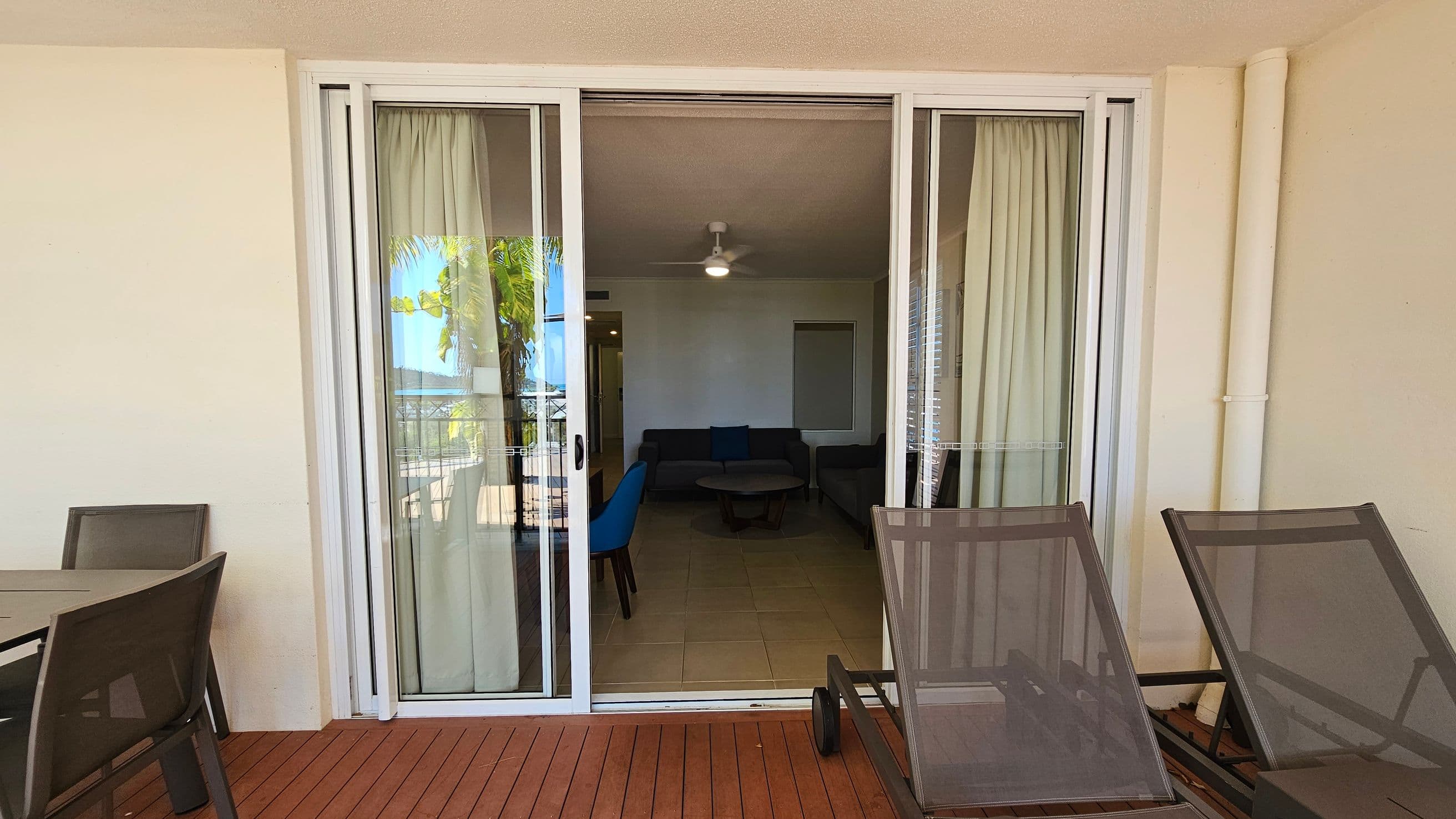 Sliding glass doors with white frames open to a living room. Inside, there's a ceiling fan and a seating area with a dark sofa and a round table. Outside, on the wooden deck, there are two lounge chairs on the right and a table with chairs on the left. A palm tree is visible through the glass, and curtains are drawn to the sides.