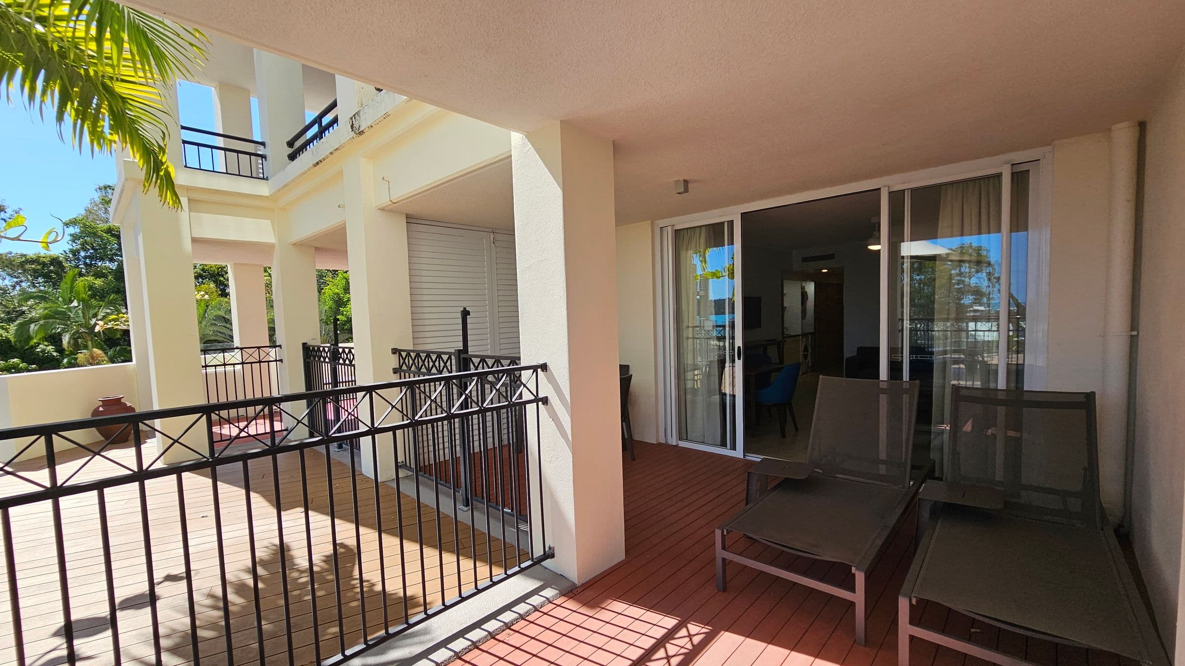 A spacious balcony with a wooden floor and cream-coloured walls. On the right, there are two dark-coloured lounge chairs in front of a sliding glass door leading inside. The left side features a black metal railing and a view of lush greenery, including palm trees. The ceiling is light-coloured, and the area is well-lit by natural light.