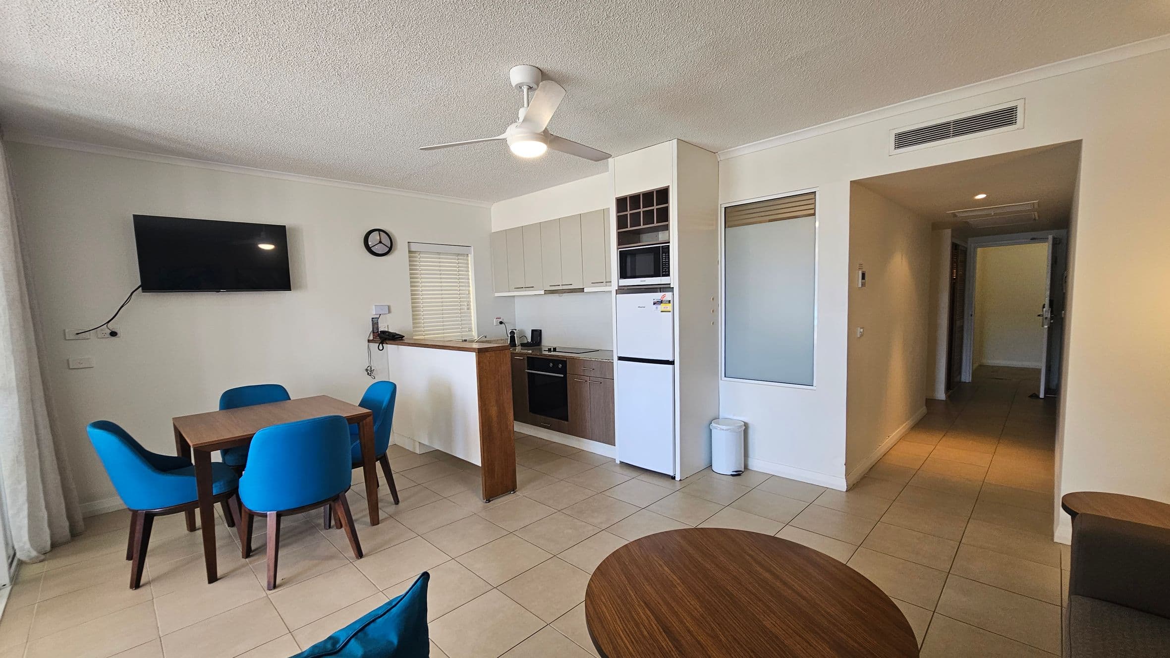 A modern living space with a tiled floor. On the left, a wall-mounted TV is above a wooden dining table with four blue chairs. The kitchen area in the centre features light-coloured cabinets, an oven, a microwave, and a fridge. A small window with blinds is above the sink. A ceiling fan with a light is overhead. On the right, a hallway leads to another room. A small round table is partially visible in the foreground.