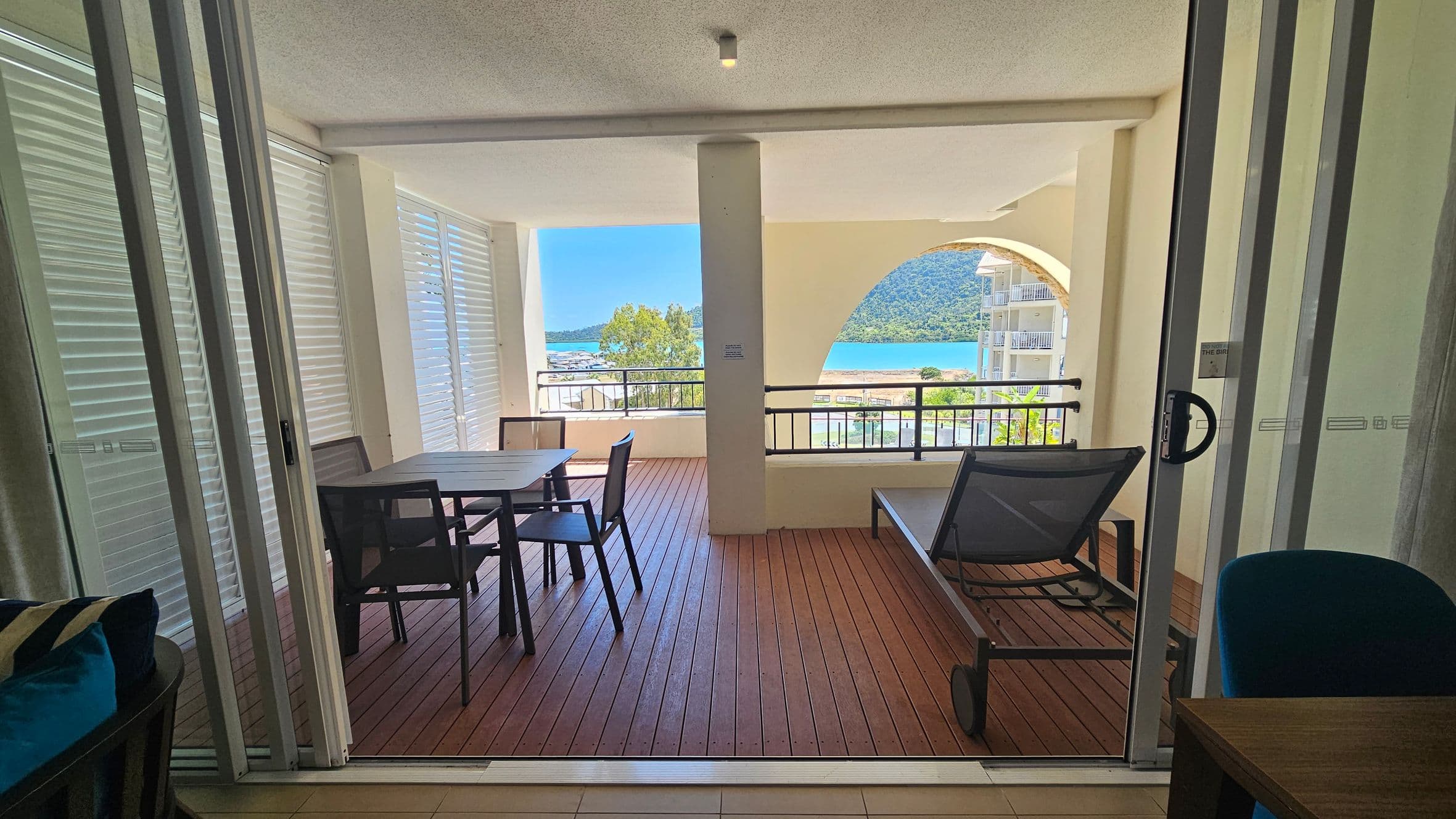 A spacious balcony with wooden flooring is visible through sliding glass doors. On the left, there's a table with four chairs. To the right, a sun lounger is positioned near the railing. The balcony is partially enclosed with white louvred panels. In the background, a scenic view of a blue body of water and a green hill is visible, framed by an arched opening. The ceiling is smooth with a small light fixture.