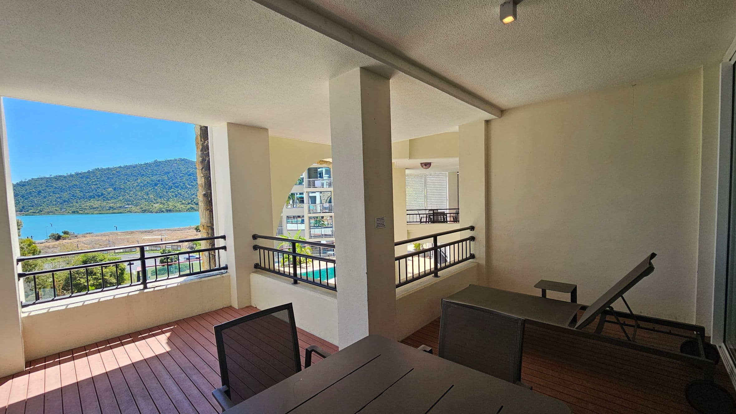 A covered balcony with a wooden floor and cream walls. On the right, there's a lounge chair and a table with chairs. The balcony is bordered by black railings. In the background, a view of a blue body of water and a green hill is visible through an open section. To the left, part of a building with balconies can be seen. The ceiling is light-coloured with a small light fixture.
