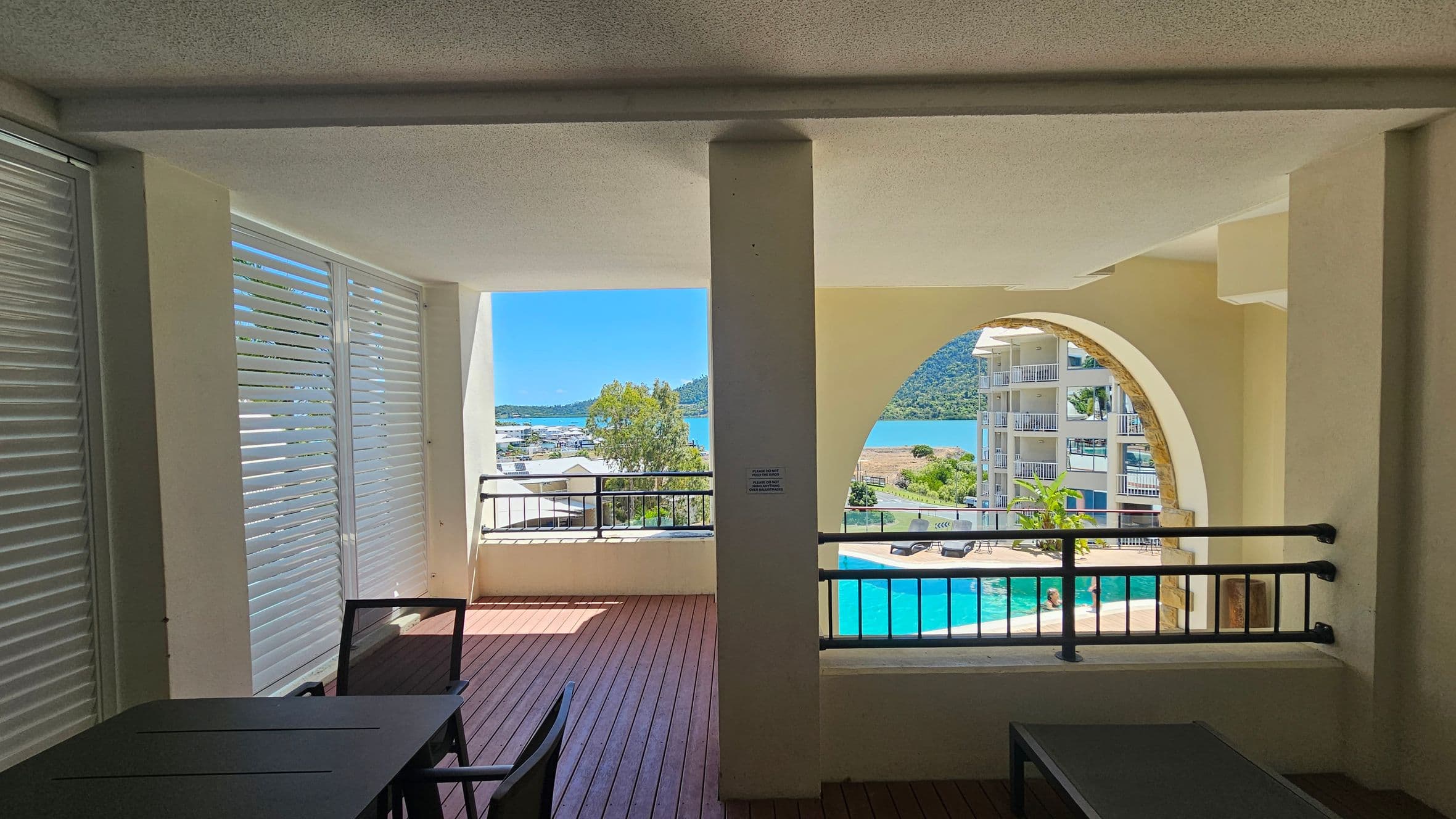 A covered balcony area with wooden flooring and a ceiling with a textured finish. On the left, there are white louvred panels. In the foreground, dark-coloured tables and chairs are visible. The middle section has a railing and a view of trees and a marina in the distance. On the right, an arched opening frames a view of a swimming pool and a multi-storey building. Beyond, there are hills and a body of water.