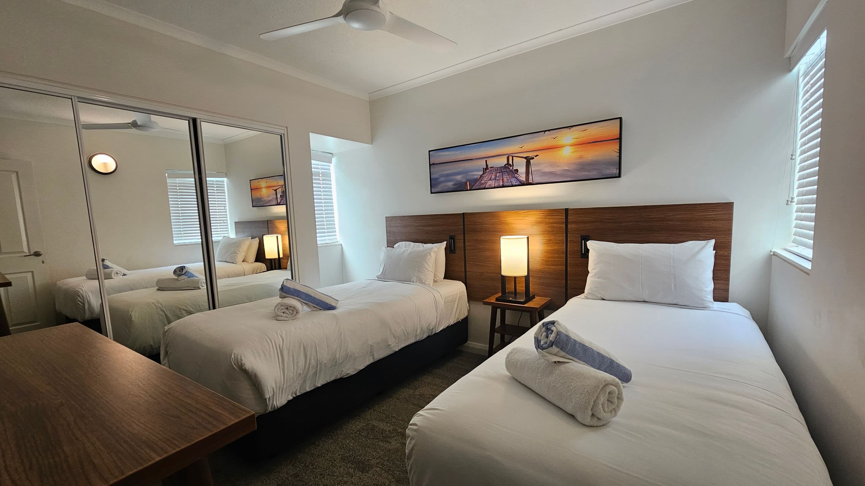 A bedroom with a large bed in the centre, covered in white linens and a towel arrangement on top. The headboard is wooden with two bedside lamps. Above the bed is a landscape picture. On the right, there are two windows with blinds and a black luggage rack. A mirrored wardrobe is also on the right. On the left, a wooden desk is partially visible. The ceiling has a white fan, and the floor is carpeted in a muted tone.