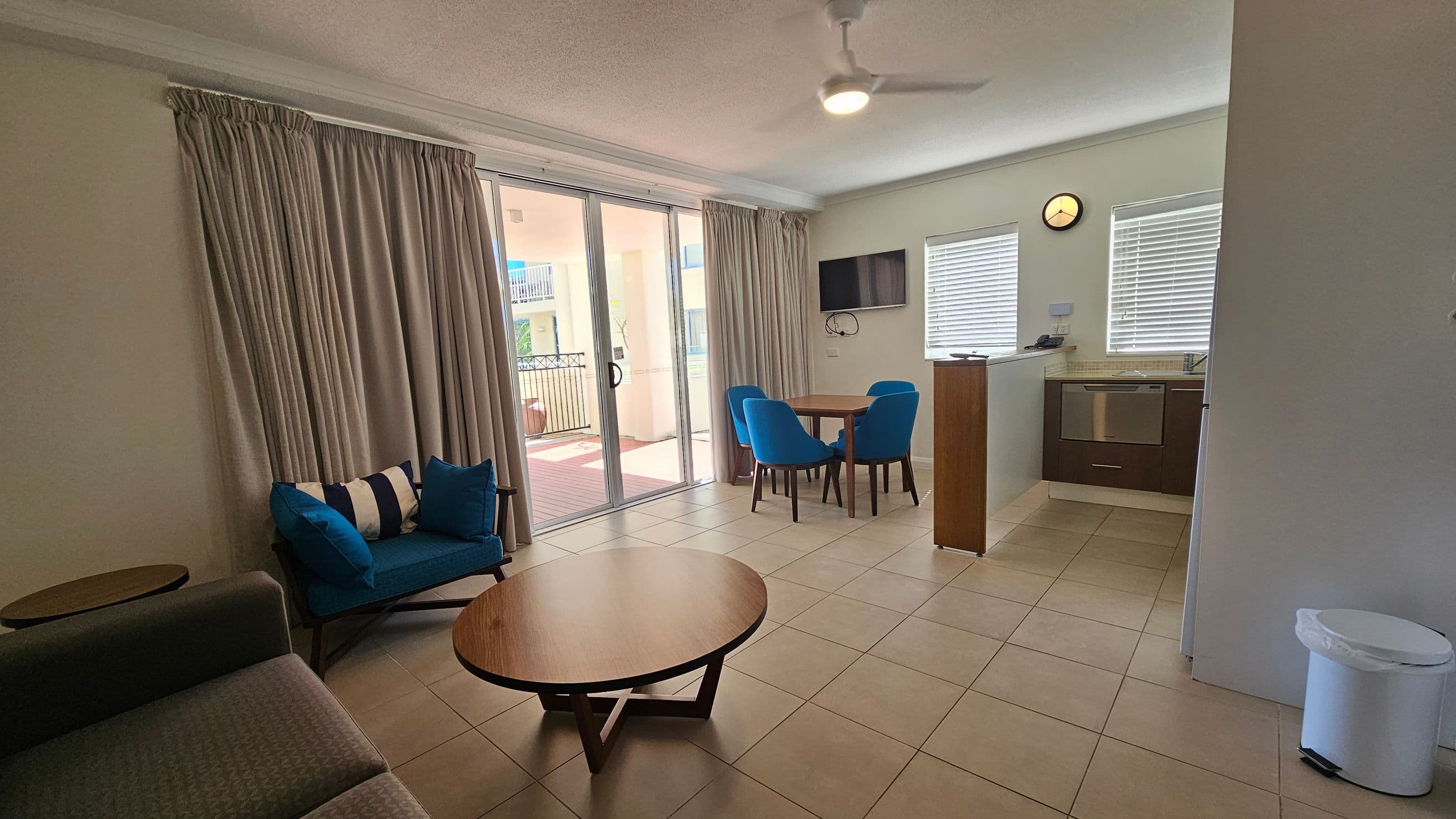 A living area with a tiled floor and a ceiling fan. On the left, there's a grey sofa and a blue armchair with a striped cushion, next to a round wooden coffee table. Large sliding glass doors with beige curtains lead to a balcony. On the right, a small dining table with four blue chairs is near a wall-mounted TV. The kitchen area has a dishwasher, two windows with blinds, and a wall clock. A white bin is in the corner.