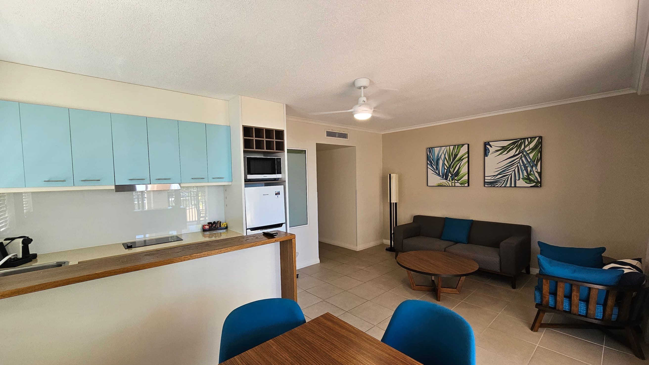 A modern living space with a kitchen on the left featuring light blue cabinets, a sink, and a stovetop. A small dining table with blue chairs is in the foreground. On the right, there's a seating area with a grey sofa, a wooden coffee table, and a chair with blue cushions. Two framed artworks with leaf designs hang on the wall. The floor is tiled, and a ceiling fan is visible above.