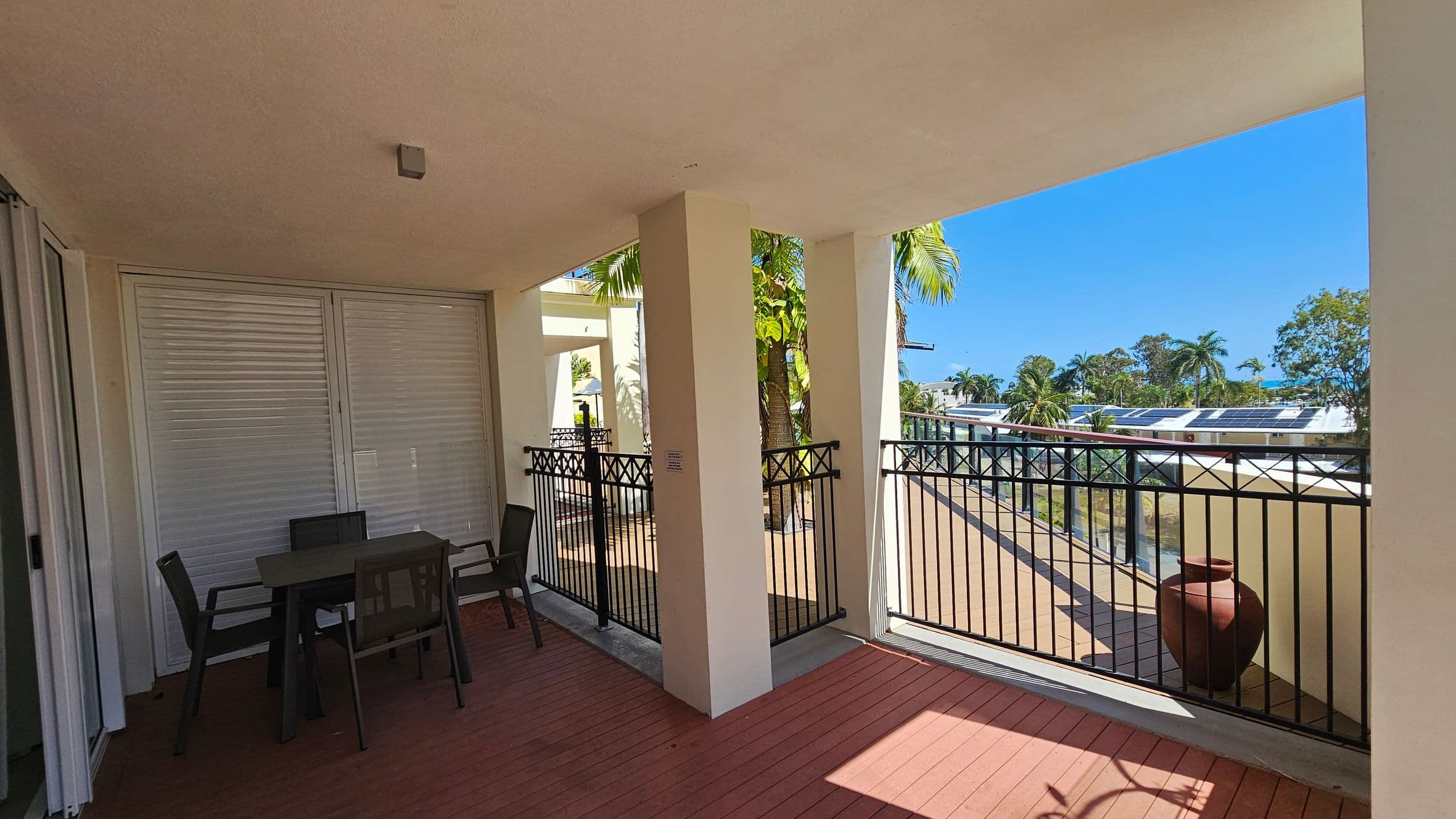 A covered balcony with a wooden floor and a light-coloured ceiling. On the left, there is a dark table with four chairs near closed shutters. In the middle, two pillars support the structure, with black metal railings between them. On the right, the balcony opens up to a view of palm trees and rooftops with solar panels. A large clay pot sits near the railing.