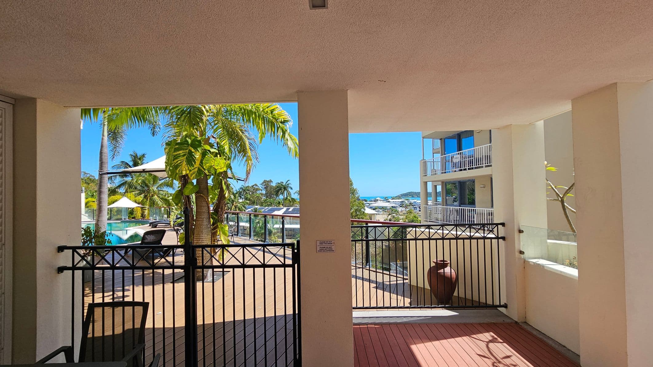 A view from a covered terrace with a textured ceiling. In the foreground, a black metal gate opens to a wooden deck. On the left, there are palm trees and a swimming pool with sun loungers and white umbrellas. On the right, a large ceramic pot sits near a glass railing. In the background, a multi-storey building with balconies overlooks a distant view of the sea and more greenery.