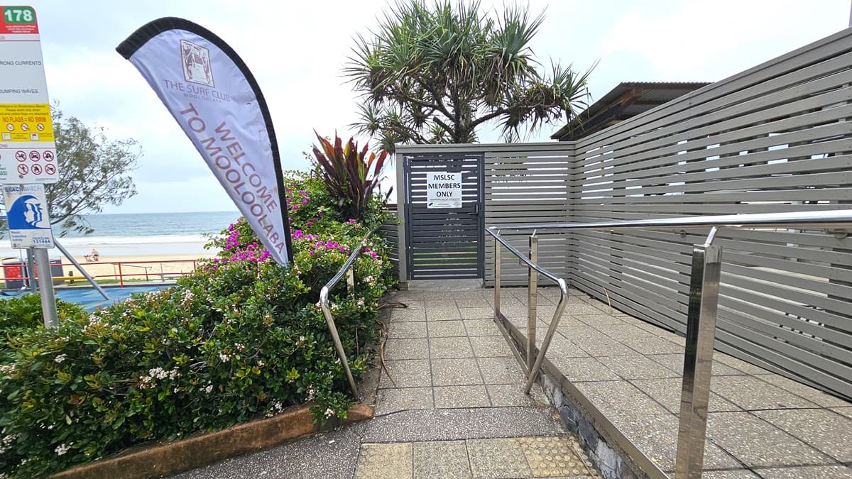 A ramp with handrails and truncated domes, bordered by landscape plants and a wooden fence. Signages affixed to a pole, including a large feather signage with the facility name. A metal louvre and gate secure the entrance, bearing a banner that reads "MSLSC MEMBERS ONLY". Palm trees are visible. An orange metal railing secures the space. The ocean is in the background.