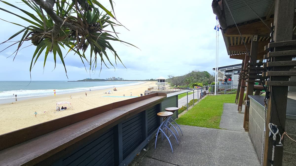 A shaded structure stands in front of a facility building, featuring wooden beams and corrugated roofing, facing the beach. Metal railing, trees, and grass border the area. Stools are arranged upfront, with rubbish bins against the wall. The beach, with a few swimmers, is visible in front, and further ahead, the patrol tower can be seen.