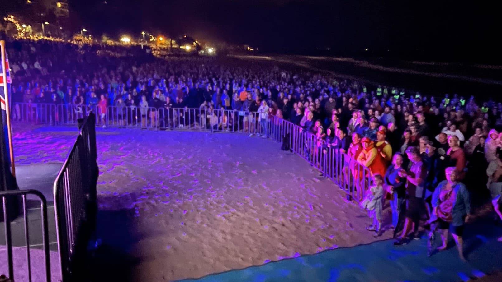 A crowd of people gathered on a sandy beach at dawn for an outdoor event. The audience is separated from a stage area by metal barriers, and the scene is illuminated with ambient stage lighting that casts a soft glow on the attendees. The backdrop features a line of trees and lights, indicating the proximity of a promenade or street parallel to the beach.