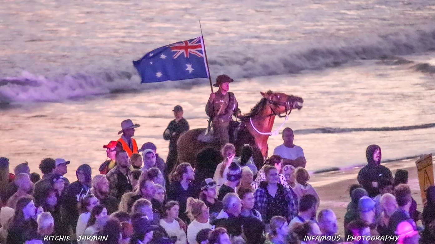 An image depicting the ANZAD DAy. A crowd of people gathered on a shore along with a man riding a horse holding the national flag.