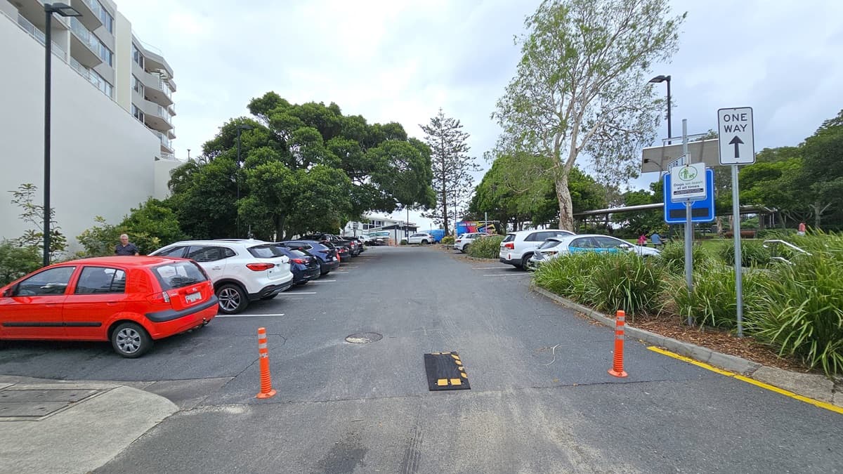 The parking area. Vehicles of various shades and models parked against the wall of a multi-storey building on the left. To the right, more vehicles parked under the shade of trees. Orange bollards placed in the middle of the road. Ornamental plants and trees occupy a concrete planter box on the right. Street signs and tall street lights are visible.