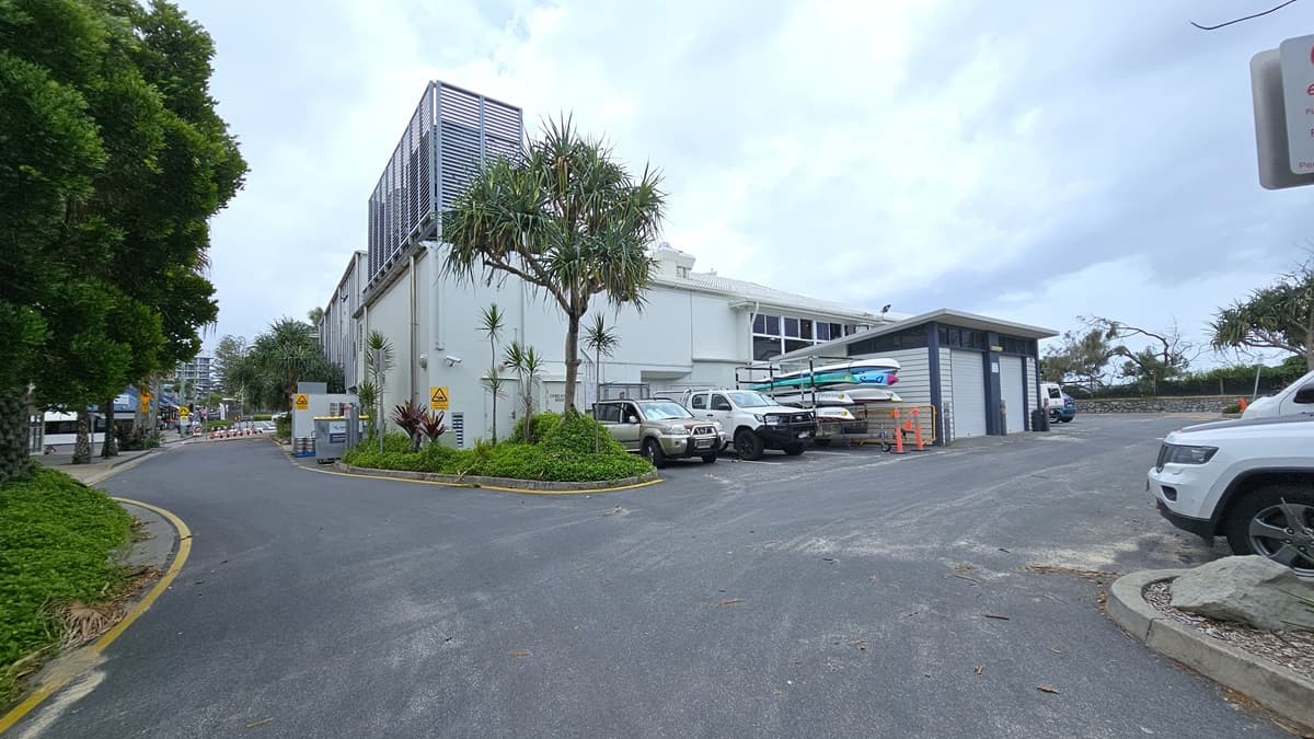 A street view showing a car park with multiple vehicles, including a white pick-up truck in the foreground. There's a small roundabout with a planted area in the centre. To the right, there's a single-storey building with signs and a petrol pump canopy. In the background, a taller modern building with vertical louvres. Landscaped plants and trees border the left. Road signs are visible.