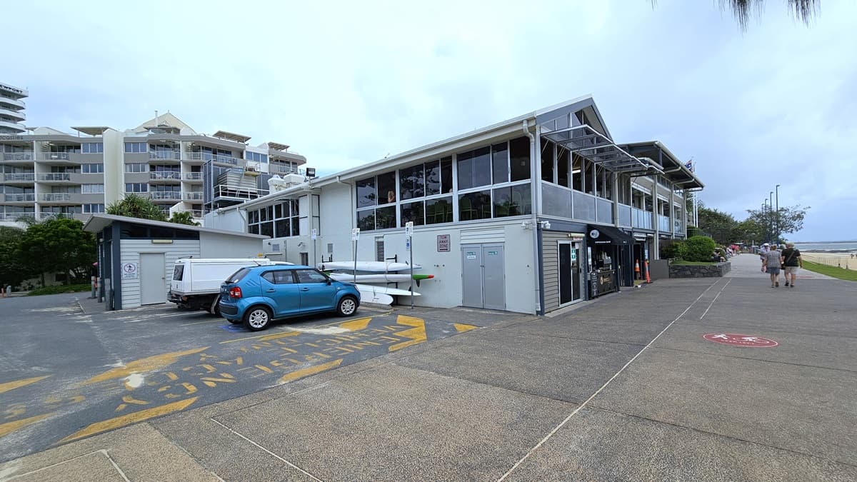 A street view showing a car park with two vehicles, including a sign for emergency parking. Adjacent is a single-storey building with a glass and concrete facade. A retail shop is on the front. To the right is a pedestrian road with railings, bordered by trees and street lights. Pedestrians are visible. A multi-storey building is in the background.
