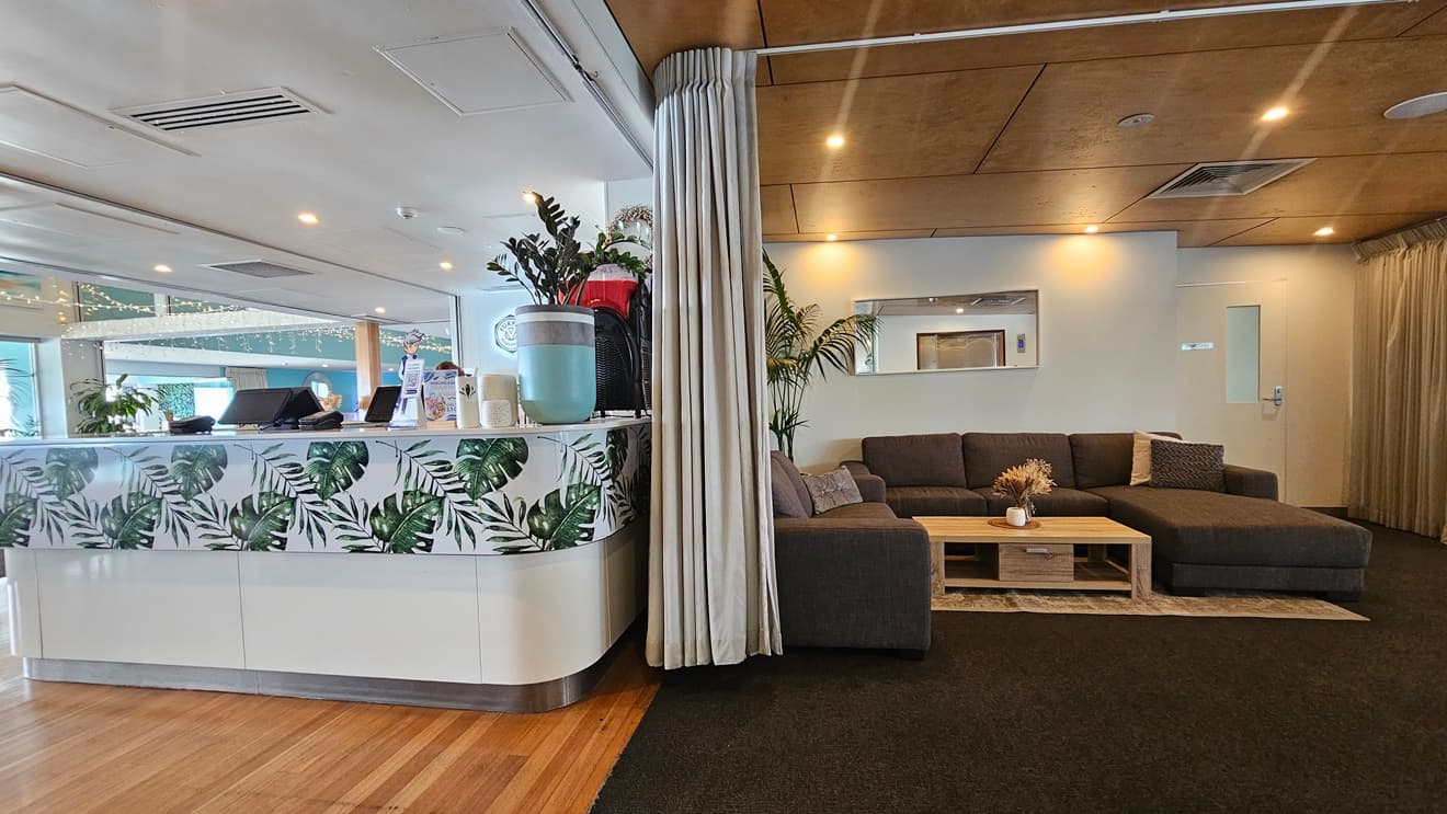 To the right is a lift, with a stairway beside it featuring handrails and truncated domes. Adjacent is a wall adorned with artificial foliage decoration. Tables and chairs are arranged in the area. The ceiling is timber with LED lights, and the floor transitions from carpet to timber.