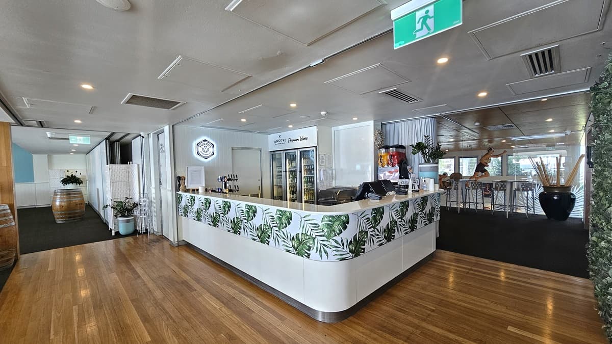 In the middle is a reception desk with a white front adorned with foliage decoration. On top are a computer, swiping machines, and indoor plant decoration. A fridge filled with beverages is positioned at the back. To the left is a carpeted area with barrel decorations. To the right, movable chairs and tables are arranged. A decoration of a man on a surfboard position is also visible. The ceiling features white LED lights and air conditioning, and the floor transitions from carpet to timber.