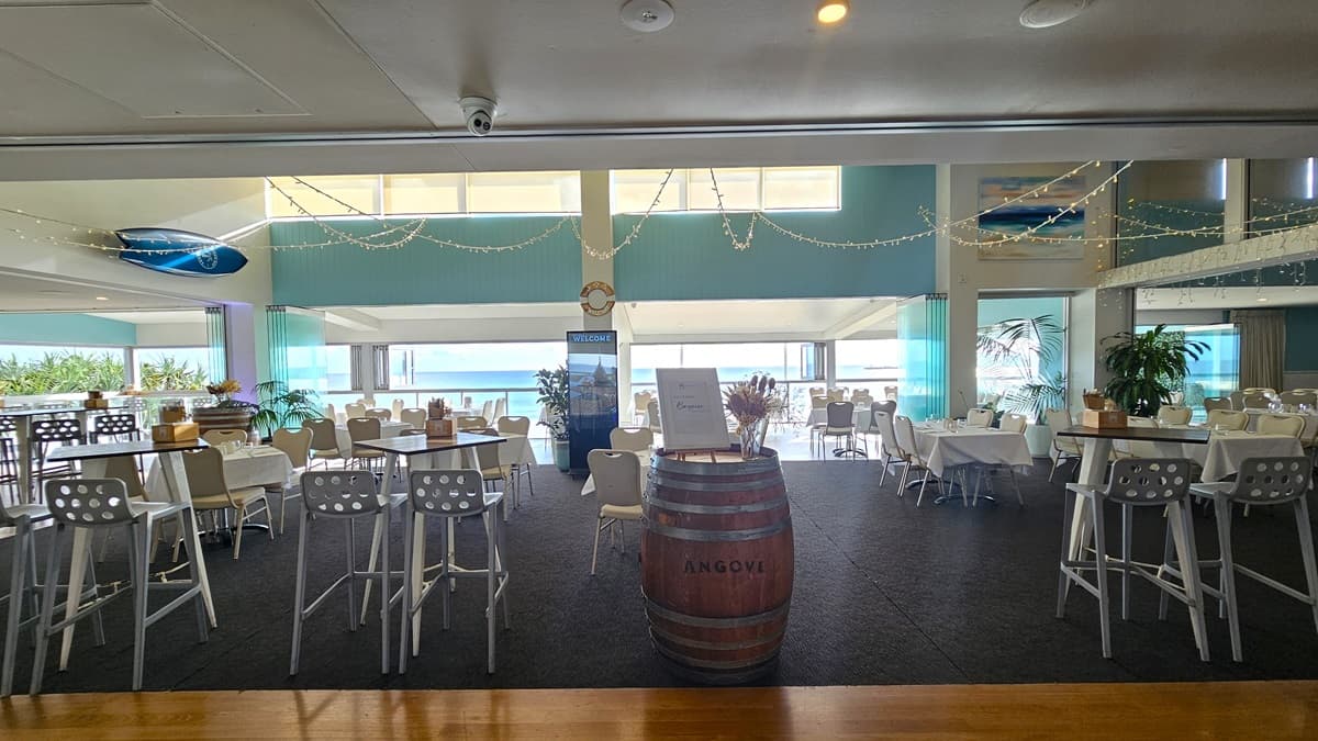 A spacious area with high-top tables and chairs. Further in, low-set chairs are arranged. Light fixtures hang overhead. The floor transitions from timber to grey carpet. A large barrel decoration sits in the middle with a vase and a banner on top. The wall features aqua paint with a surfboard decoration. A large retractable banner is positioned in the middle. The ceiling features white LED lights and air conditioning.