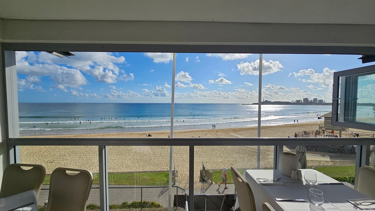 A beachside restaurant with arranged tables and chairs overlooking the beach. Glass walls secure the space. Metal railings and landscape plants border the road below. Swimmers populate the beach under a sunny sky with white cloud formations. A shaded structure is also visible.
