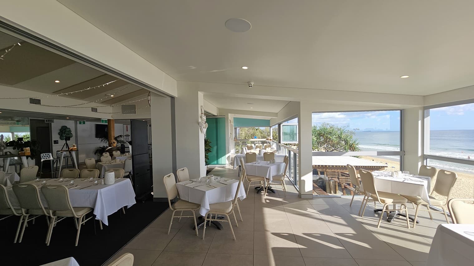 Tables and chairs are neatly arranged, each with a white tablecloth and utensils. Indoor plants adorn the space. The floor transitions from carpet to white tiles. A glass wall secures the area on the right. The wall and ceiling are white with aqua accents.