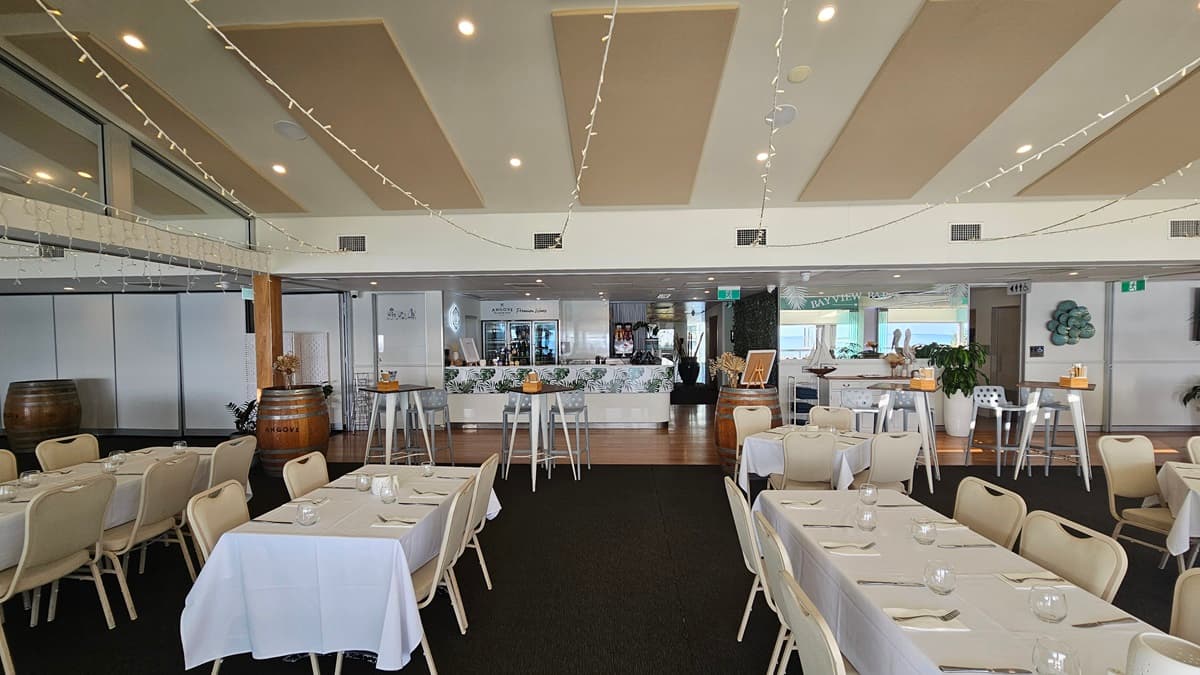 Tables and chairs are neatly set with white tablecloths and utensils. Indoor plants and barrel decorations add to the ambiance. Further in is a reception counter adorned with foliage. The floor transitions from carpet to white tiles. Walls and ceilings are white with cream accents. LED lights and air conditioning are overhead, with hanging light fixtures.