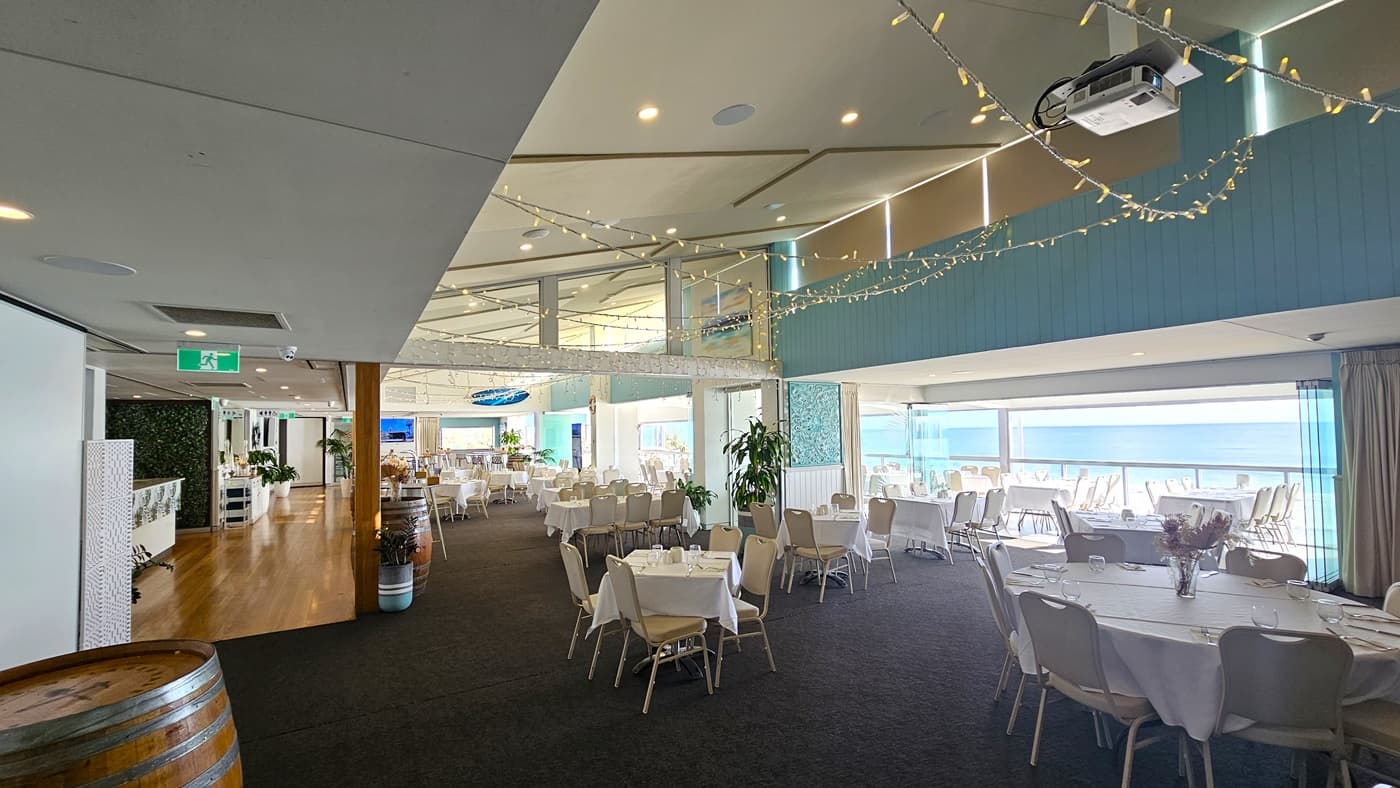 Tables and chairs are neatly arranged with white tablecloths and utensils. Indoor plants and barrel decorations enhance the atmosphere. A reception counter adorned with foliage stands further in. The walls and ceilings feature white with aqua accents. Projector machines and LED lights hang from the ceiling, alongside air conditioning and hanging light fixtures. The floor transitions from carpet to timber.