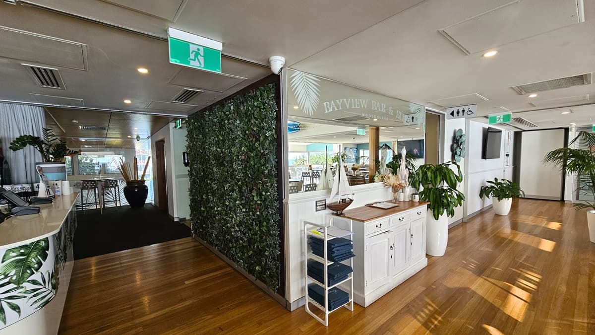 On the left stands a reception counter adorned with foliage. The right wall is decorated with artificial foliage. A mirror and cabinet with decorations are on the right. A rack holding menu books is on the left. Lights hang from the ceiling, alongside air conditioning and hanging light fixtures. The floor transitions from carpet to timber. Directional signages, including a flatscreen TV, are affixed to the upper wall. Indoor plants decorate the space.