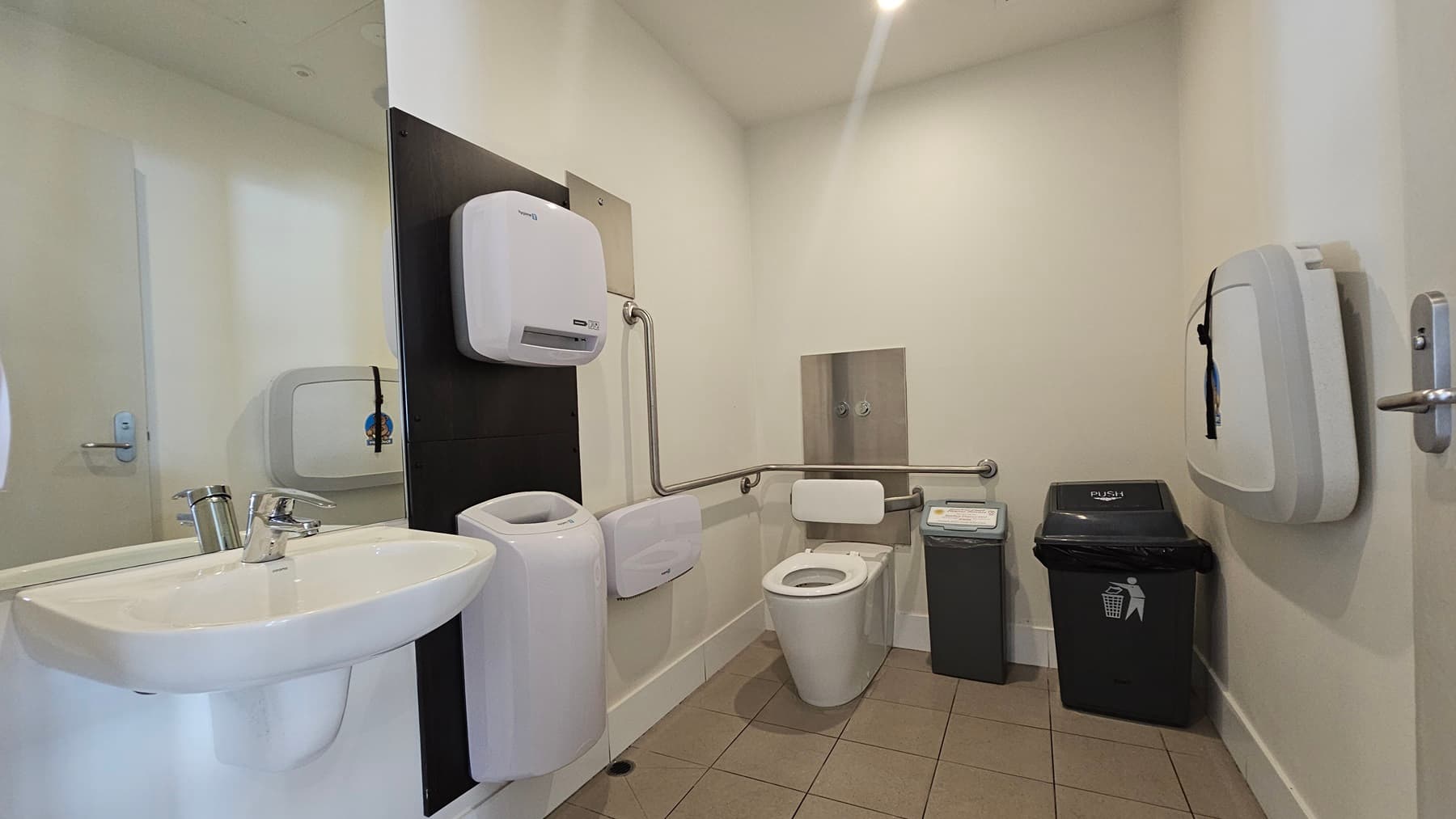 The accessible bathroom includes a toilet bowl with handrails in the corner, accompanied by a tissue dispenser and rubbish bins. On the left, there's a hand basin with a wall-mounted mirror. To the right, a wall-mounted changing table is present. The floor is tiled in light brown, while the walls are adorned with white and cream tiles. The ceiling is white, illuminated by fluorescent lights.