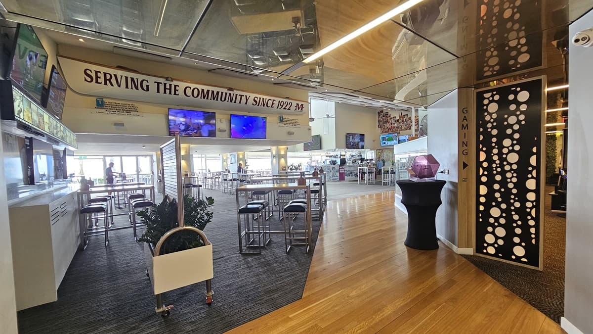 To the left is an area with carpet. The bar is in the middle, surrounded by high tables and chairs. An indoor plant decorates the left. To the right is an area with a timber floor and a wooden partition. The reception desk is further ahead. A Keno machine and a free-standing banner are in the middle. Posters, photos, flat-screen TVs, and wall decorations adorn the upper wall. A replica of a man on a surfboard is atop the wooden partition. The ceiling is white with LED lights and a ceiling fan.