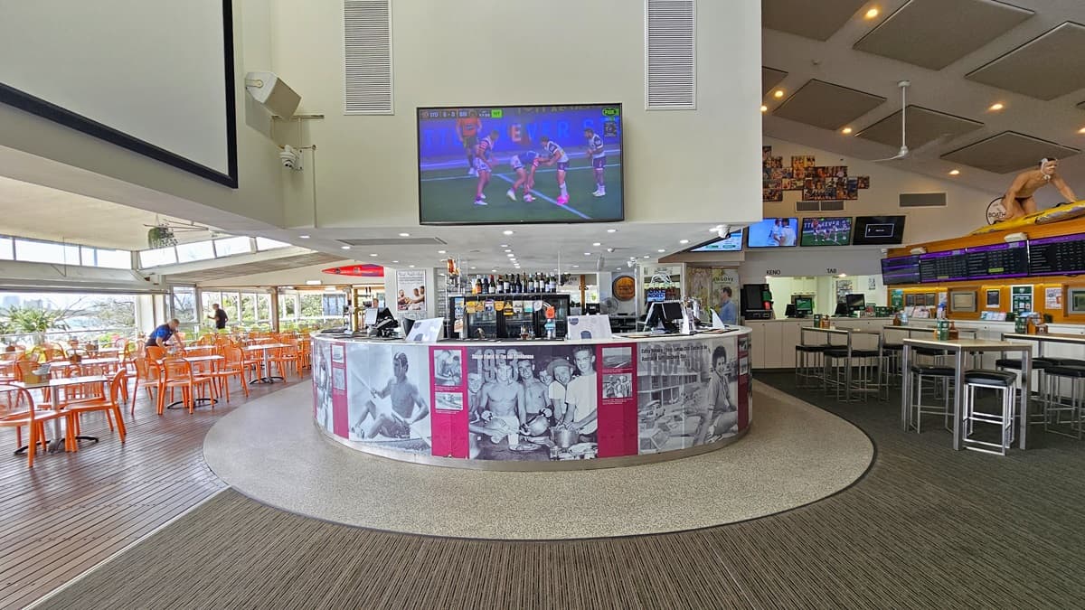 The bar has a circular counter with large posters on the facade. Behind it, a display rack showcases wines and liquors. A big flatscreen TV is mounted overhead. On the right, there's a seating area with high tables and chairs, adorned with more flatscreen TVs, posters, and a surfing-themed decoration. On the left, a large projection board is present. The floor transitions from timber to carpet and tiles. The ceiling has rectangular accents with LED lights and ceiling fans.