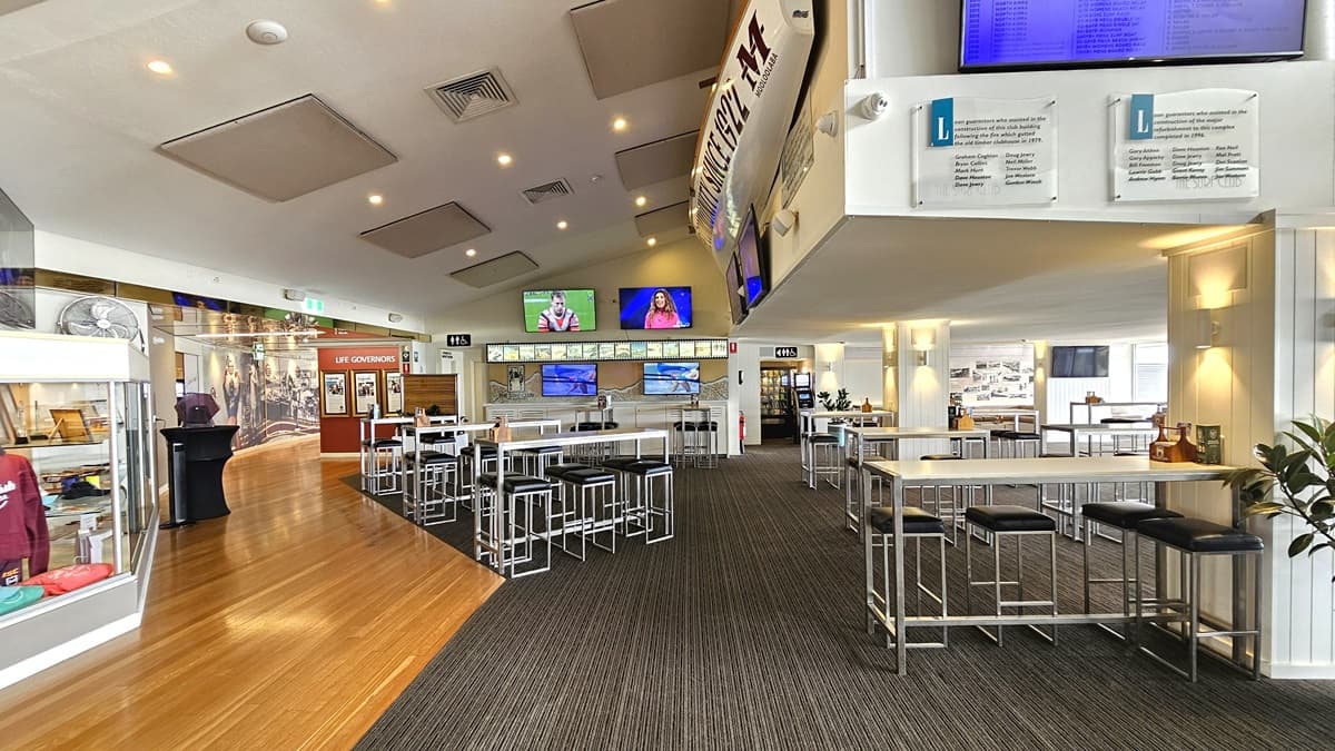 A seating area with high tables and chairs, featuring flatscreen TVs. A signage for accessible toilets is affixed to the middle walls. On the left, a small retail area displays clothing for sale. Wall fans and decorations are visible. The floor transitions from timber to carpet. Posters and signages surround the space. The ceiling has rectangular accents with LED lights and ceiling fans.
