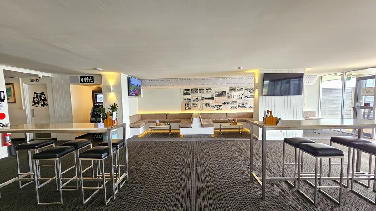 In the middle, there are couches with coffee tables. Behind, the wall is adorned with posters and photos. To the left is an accessible toilet, marked by a signage. An ATM is positioned in the corner. Flatscreen TVs and fire extinguishers are mounted to the wall. The ceiling is white with LED lights. High tables and chairs are arranged in the foreground. The floor has grey carpet.