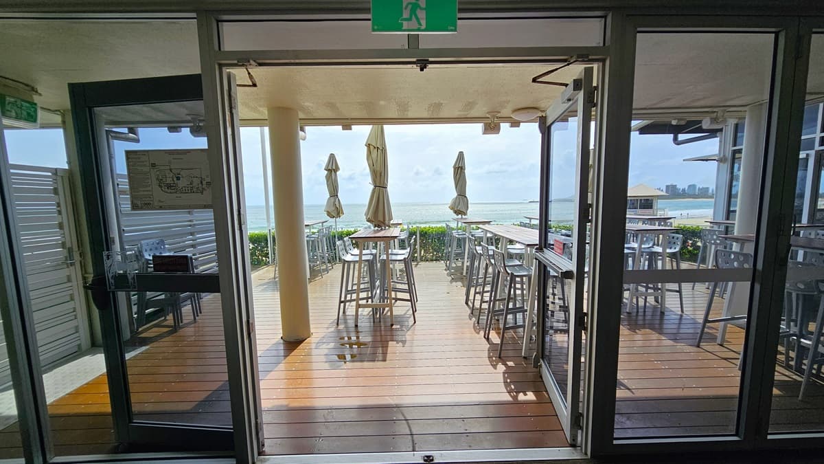 An outward-opening glass door leads to an open space with beach views. High tables and chairs are arranged, with closed large umbrella shades. Trimmed plants border the front. The floor has timber, and an exit sign is affixed to the door.