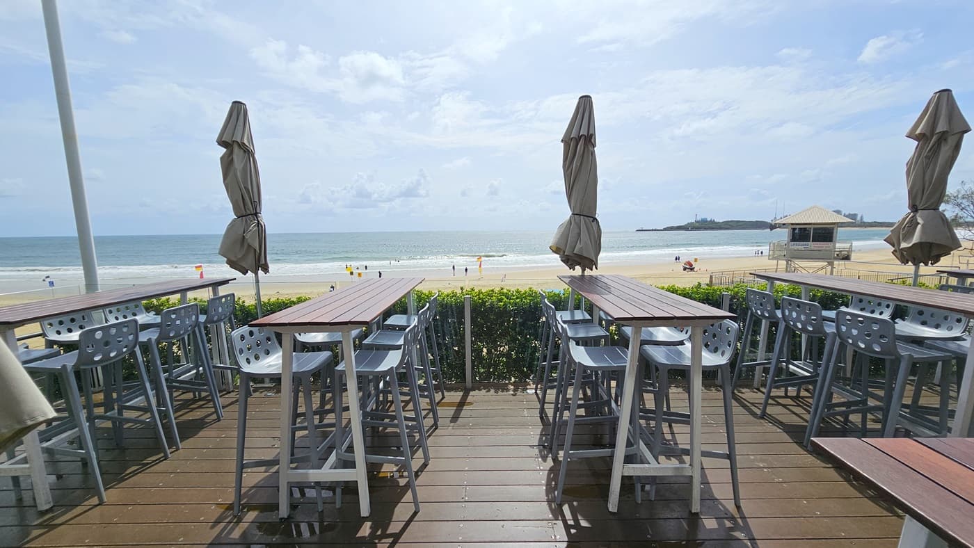 An open space overlooking the beach. Rows of high wooden tables and grey chairs are arranged, each with closed large umbrella shades. Trimmed plants border the front. To the far left is a shaded structure with a viewing deck. The floor is timber.