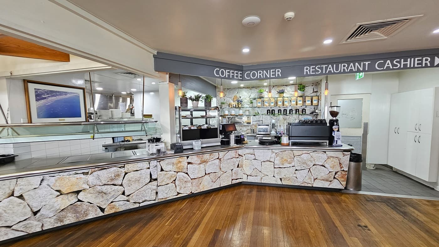 A lengthy reception area with a stone front decoration. Food display racks and condiments are atop. A utensil rack is fixed to the wall. Signages for "Coffee Corner" and "Restaurant Cashier" are near the ceiling. Cabinets are to the right. The floor is timber, and the ceiling is white with LED lights and air conditioning.