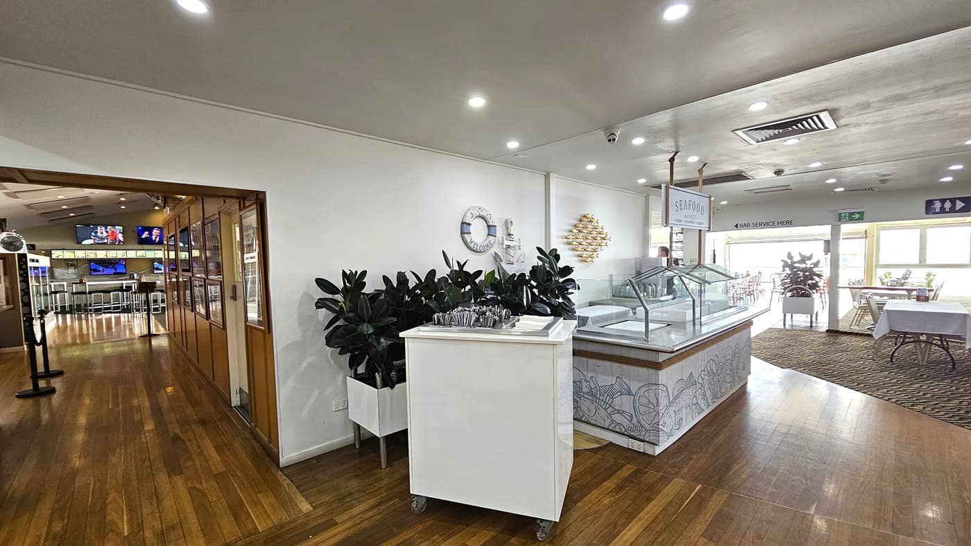 A seafood counter adorned with seafood decorations and food racks. Adjacent, a utensil rack stands beside a planter box with indoor plants. To the left, a corridor with timber flooring leads to the entrance and more seating. On the right, a carpeted area with arranged seating. Flatscreen TVs and directional signages are visible. The ceiling is white with LED lights and air conditioning.