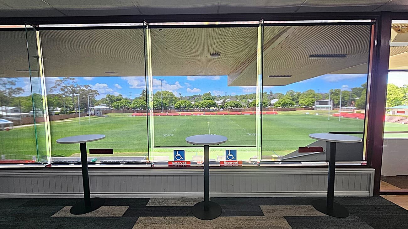 A view from inside a building with large glass windows overlooking a green sports field. Three tall round tables with black bases are positioned in front of the windows. The floor is carpeted in a dark pattern. Outside, there are trees and a few small structures in the background. Two signs on the window indicate reserved seating for accessibility. The ceiling inside is light-coloured with a textured pattern.