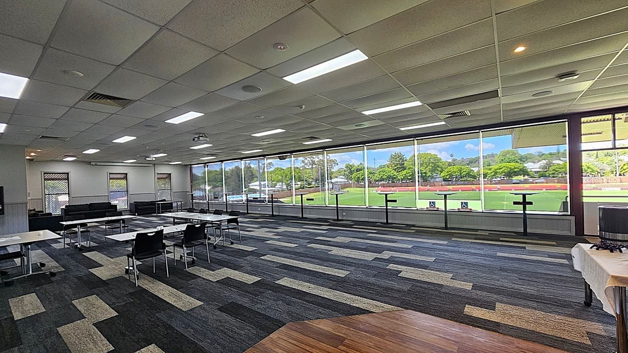 A spacious room with a patterned carpet in shades of grey and green. On the left, there are several tables and chairs arranged in a meeting setup. The ceiling is fitted with fluorescent lights and air vents. The right side features large windows offering a view of a green sports field and trees outside. In the foreground, a table with a white cloth is visible.