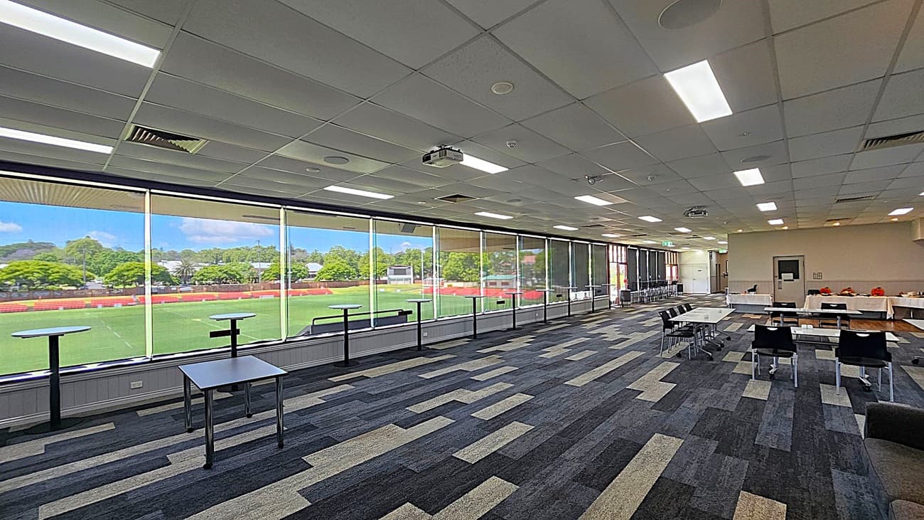 A spacious room with large floor-to-ceiling windows on the left, offering a view of a green sports field and trees outside. The room has a patterned carpet in shades of grey and beige. On the right, there are tables and chairs arranged in rows, with some tables covered in white cloths. The ceiling is fitted with fluorescent lights and a projector.